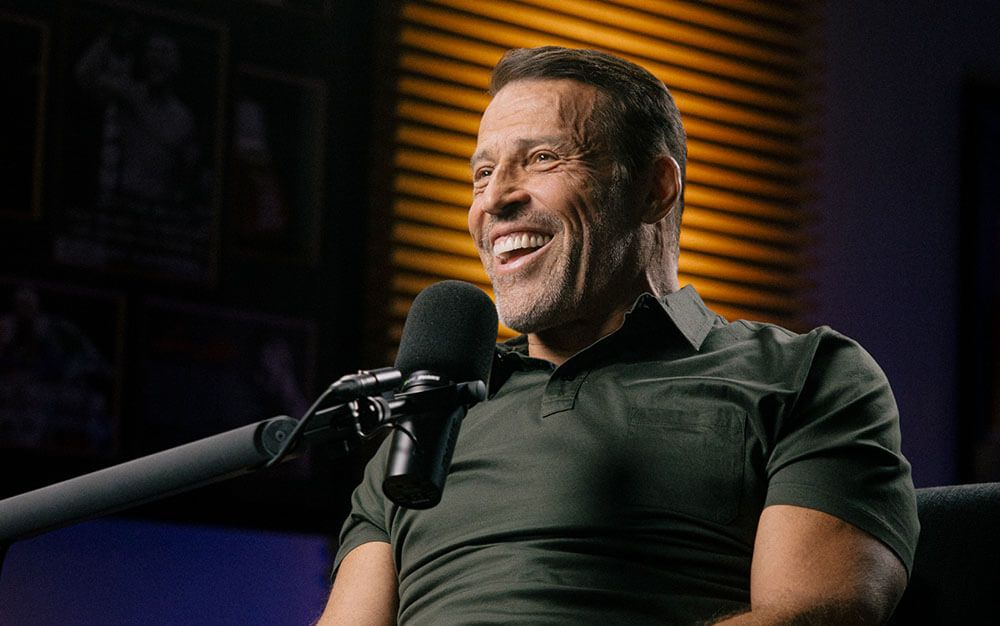 Tony is sitting in a suit and tie holding a microphone.