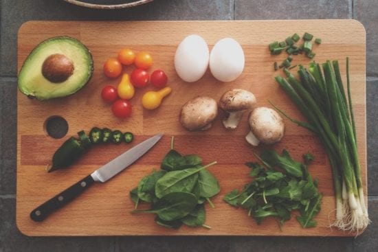a wooden cutting board topped with avocado , eggs , mushrooms , spinach , and green onions .