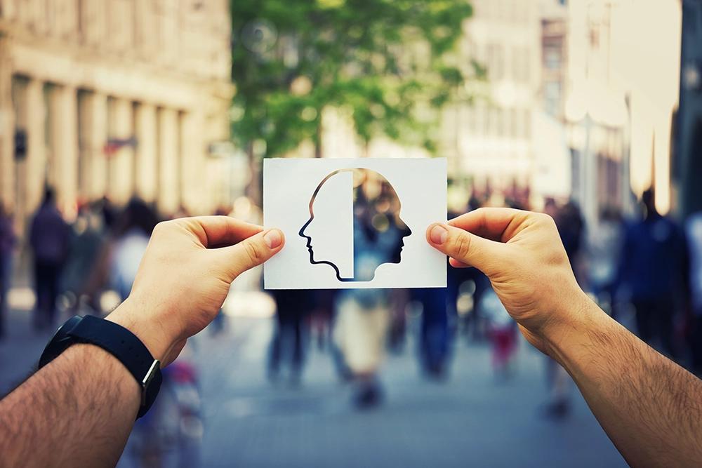 Hands hold a paper cutout of two intertwined head silhouettes, revealing a blurry city street behind.