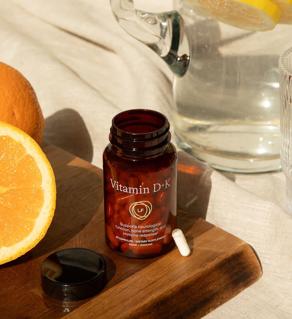 An open amber bottle of Vitamin D+K supplements, with one white capsule, next to sliced oranges and a pitcher of lemon water on a wooden surface.