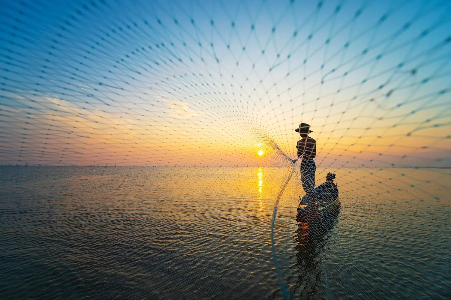 Silhouette of a fisherman casting a net from a boat at sunset, viewed through the net.
