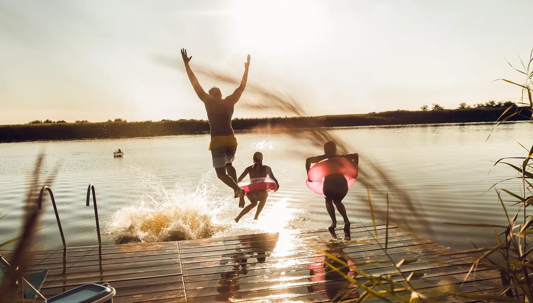 Three silhouetted people jump into a lake from a dock at sunset.