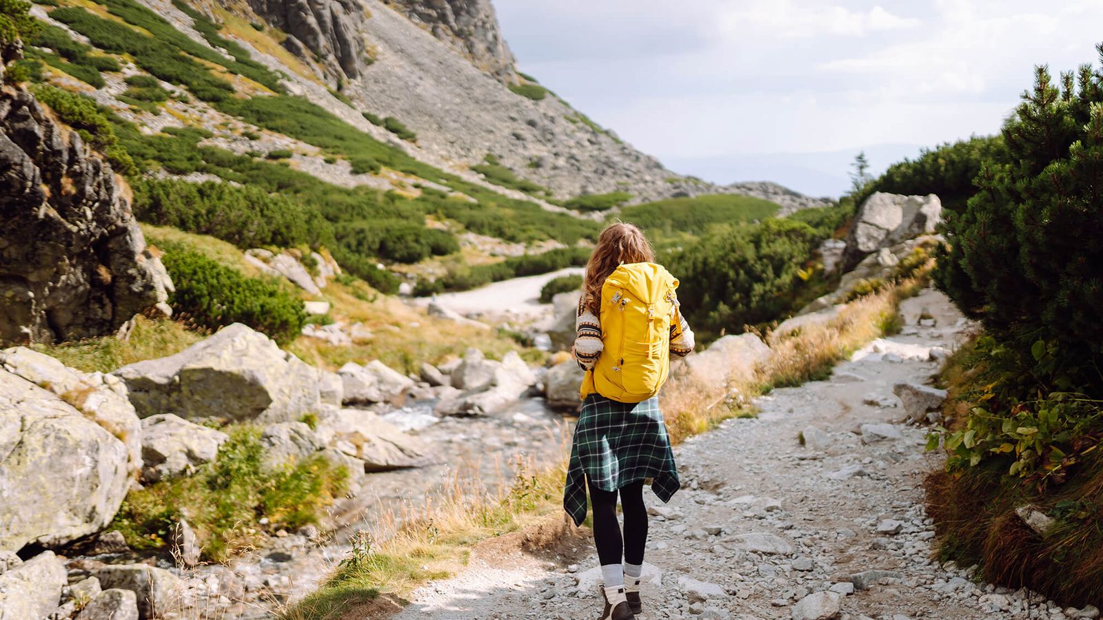 A person with a yellow backpack hikes on a rocky mountain path beside a stream.
