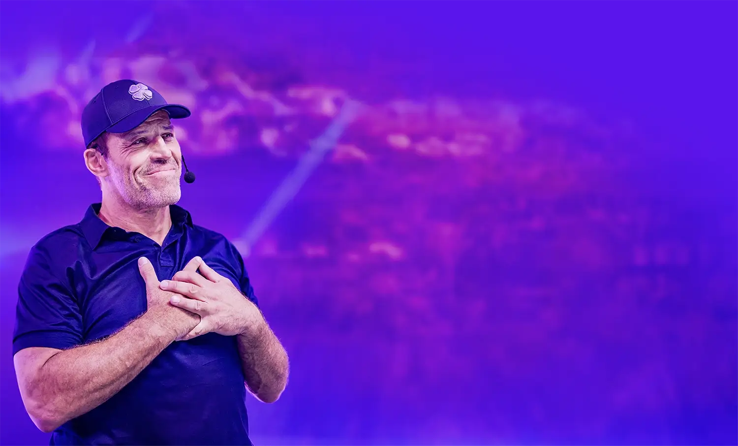 Tony Robbins, wearing a blue polo and cap, smiles with his hands clasped over his chest on a purple-lit stage.