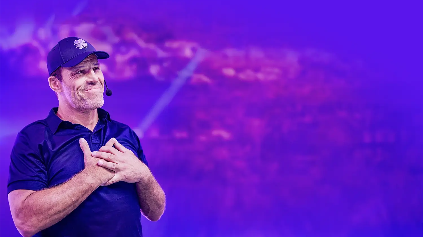 Tony Robbins, wearing a blue polo and cap, smiles with his hands clasped over his chest on a purple-lit stage.