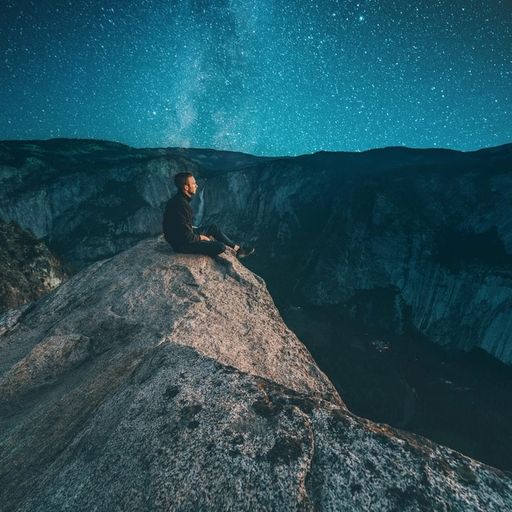 a man is sitting on the edge of a cliff at night under a starry sky .