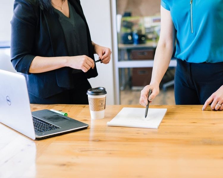 two women are standing at a table with a laptop and a cup of coffee .