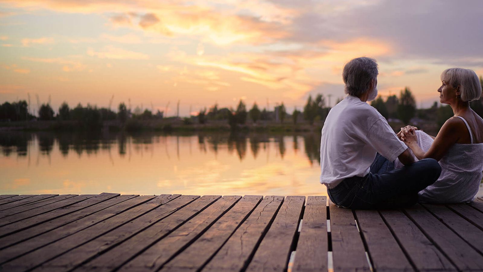 Older couple holding hands on a wooden dock overlooking a lake at sunset.