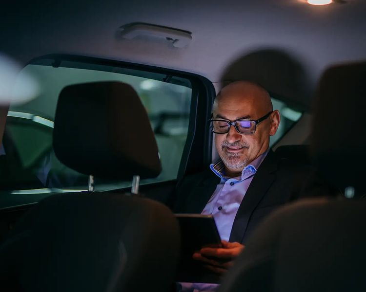 A smiling, bald man wearing glasses sits in the back of a car, illuminated by a device in his hands.