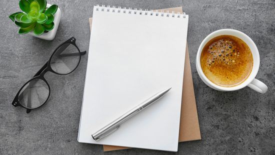 a notebook , pen , glasses and a cup of coffee on a table .
