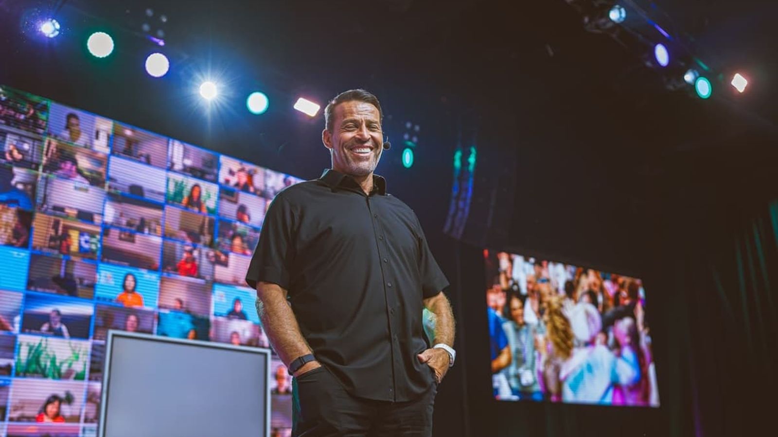 A smiling man stands on a stage with large screens displaying a grid of virtual attendees and a live crowd.