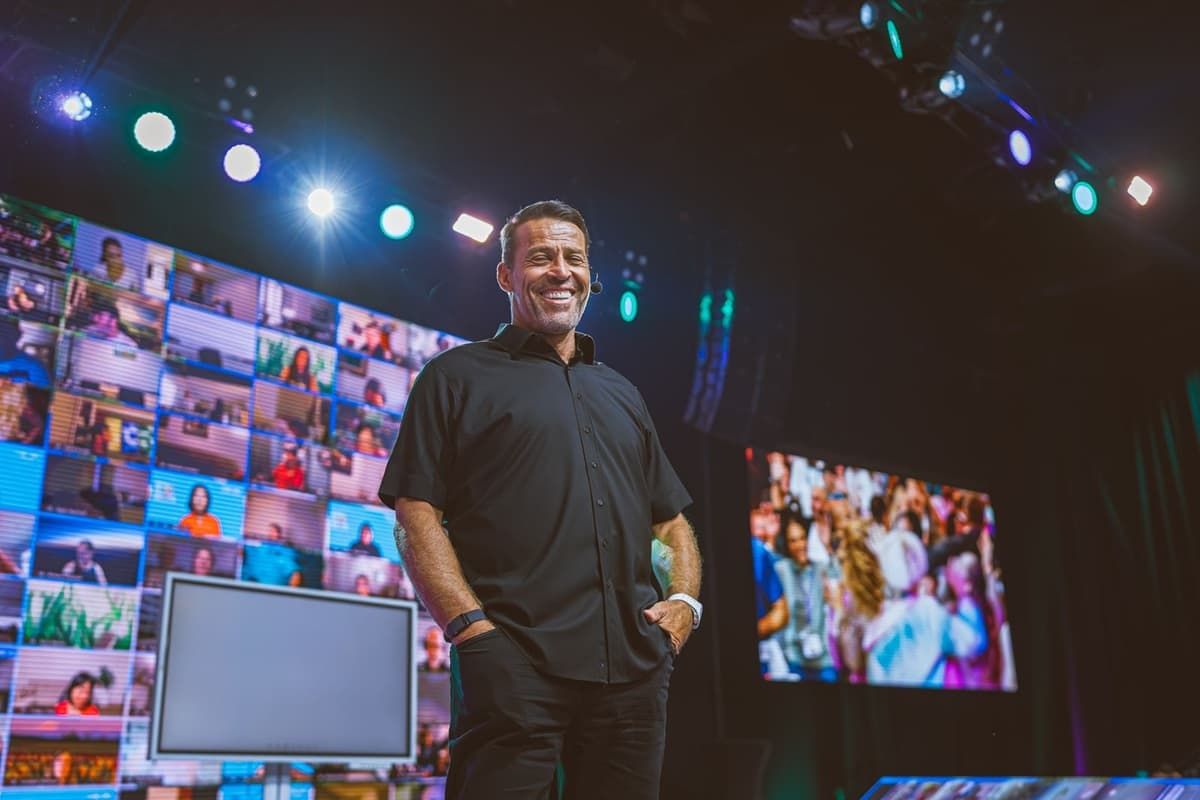 Tony Robbins smiles on stage, flanked by screens showing virtual attendees and a live crowd.