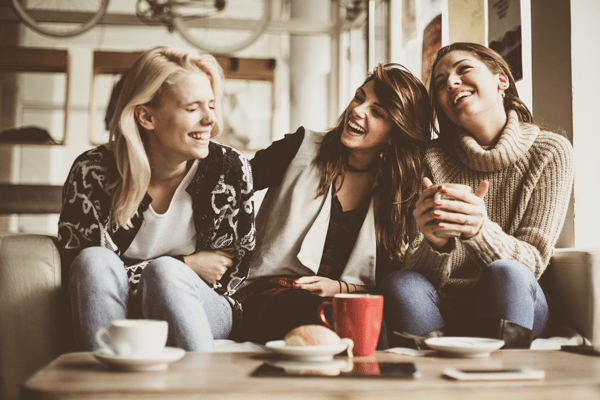 three women sit on a couch laughing and drinking coffee