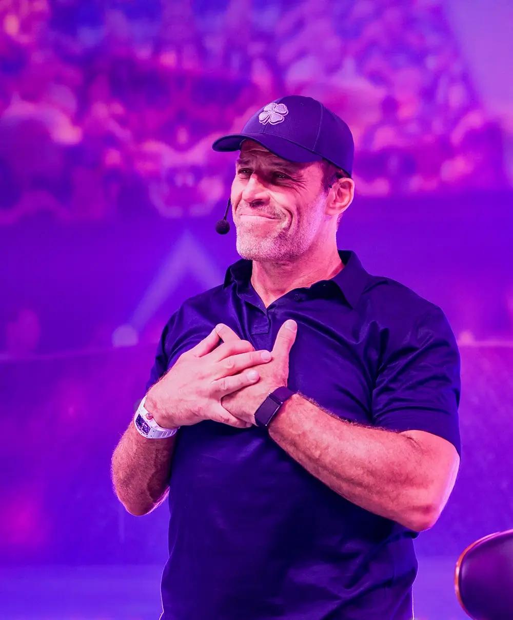 Tony Robbins, wearing a cap and lavalier mic, holds his hands over his chest with a heartfelt smile against a purple background.
