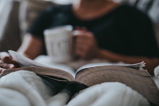 a person is sitting on a couch reading a book and drinking coffee .