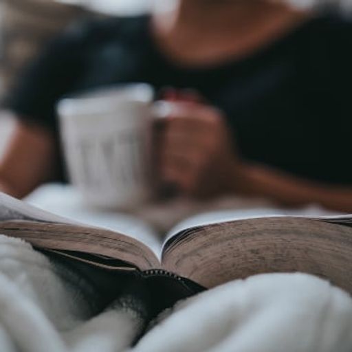 a person is sitting on a couch reading a book and drinking coffee .