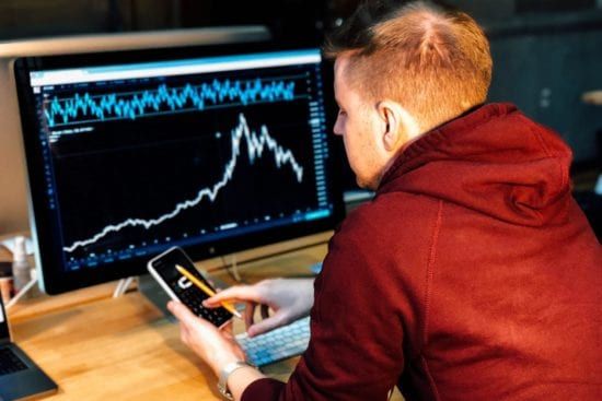 a man is sitting at a desk looking at a computer screen while holding a cell phone .