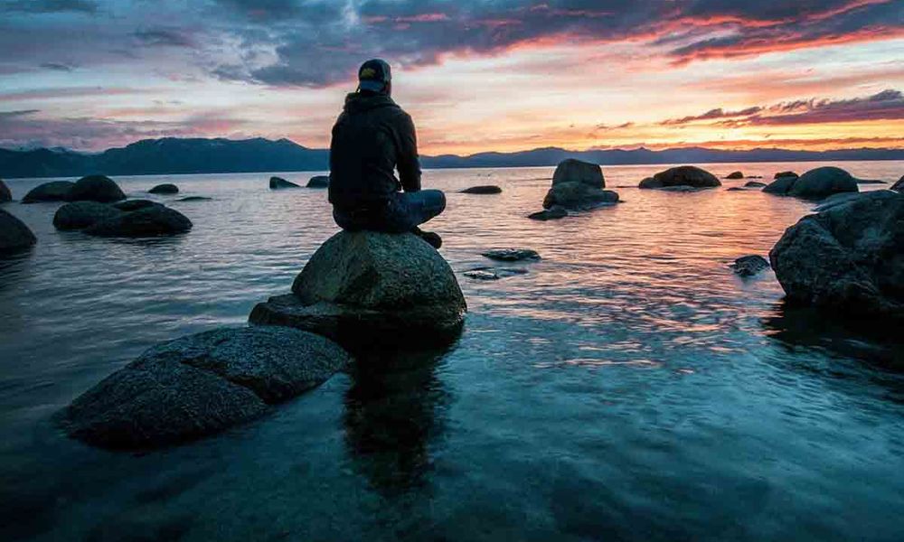 Person silhouetted on a rock in a lake, looking at a colorful sunset.
