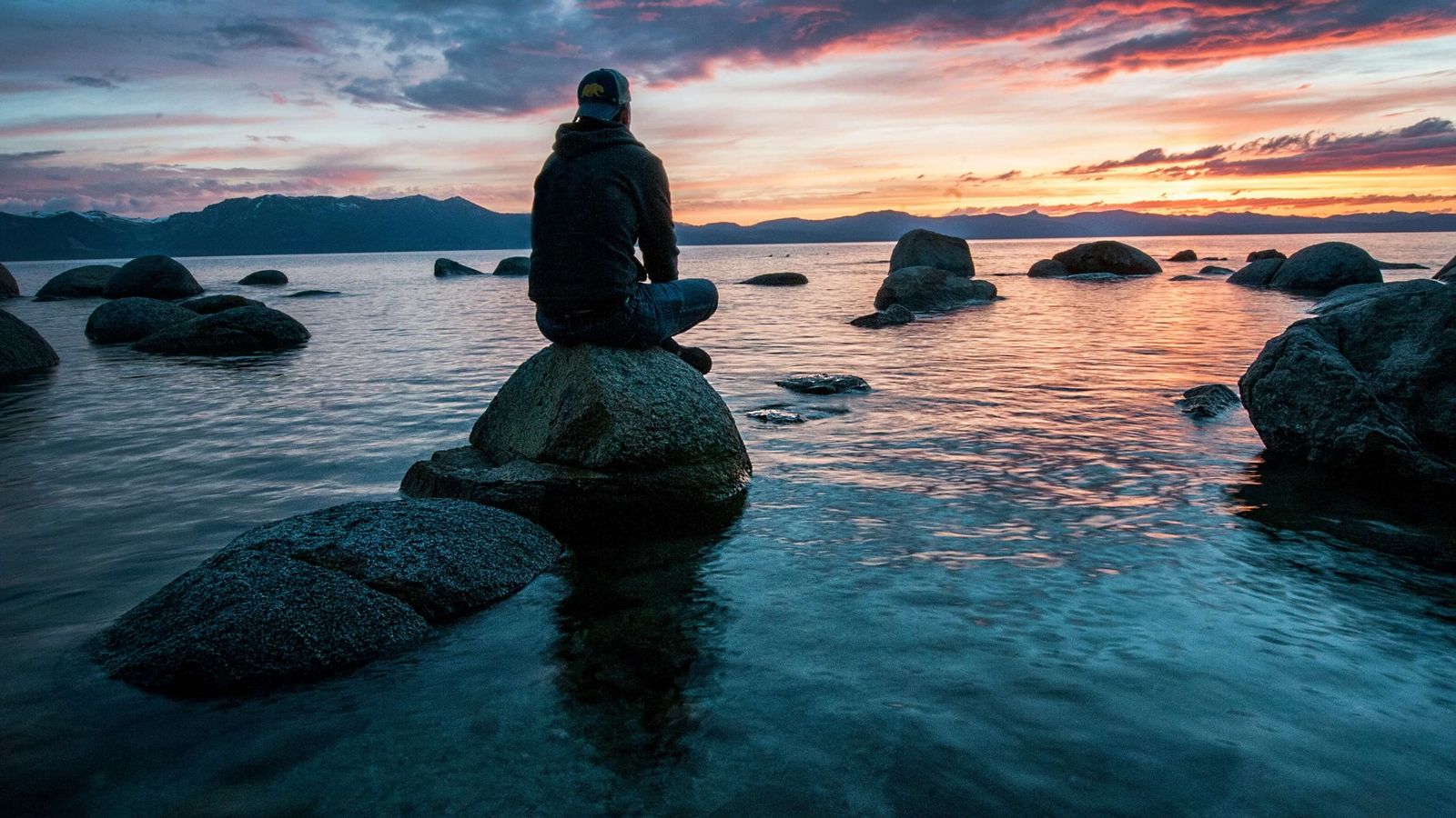a man is sitting on a rock overlooking a lake at sunset .