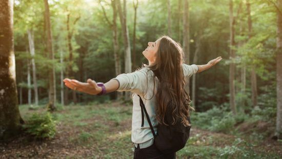 a woman with a backpack is standing in the woods with her arms outstretched .