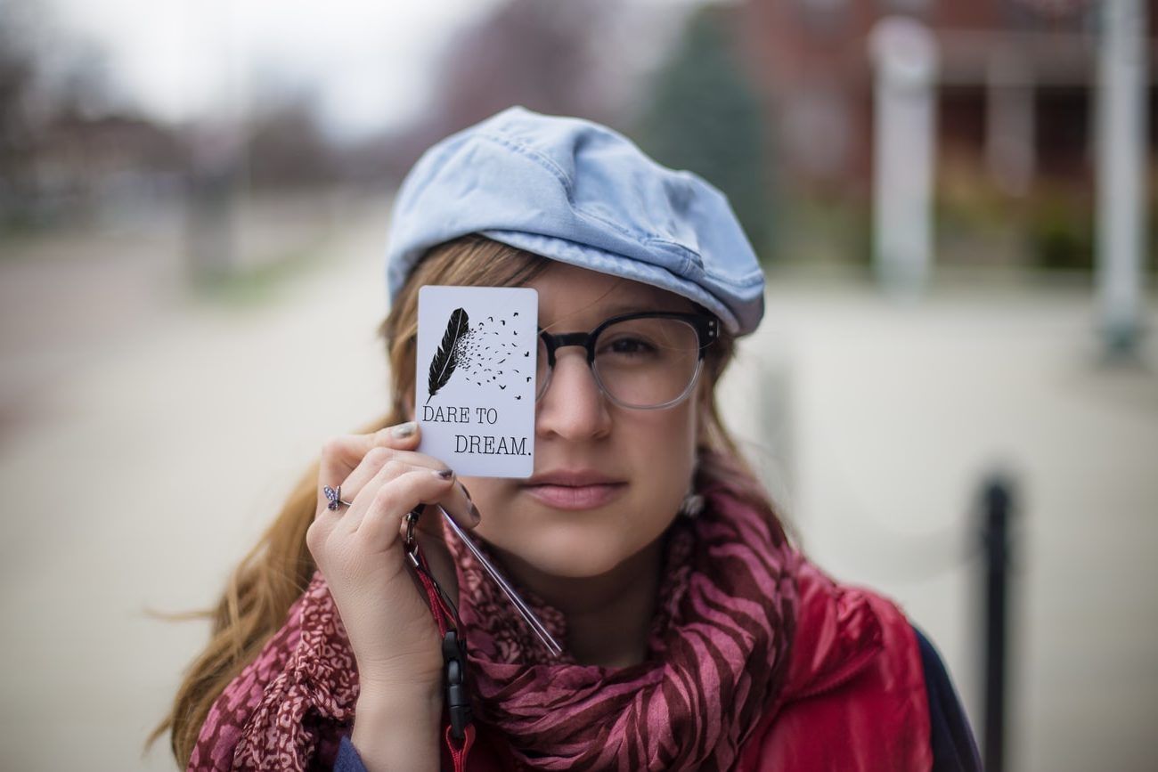 a woman wearing glasses and a hat is holding a piece of paper in front of her eye .