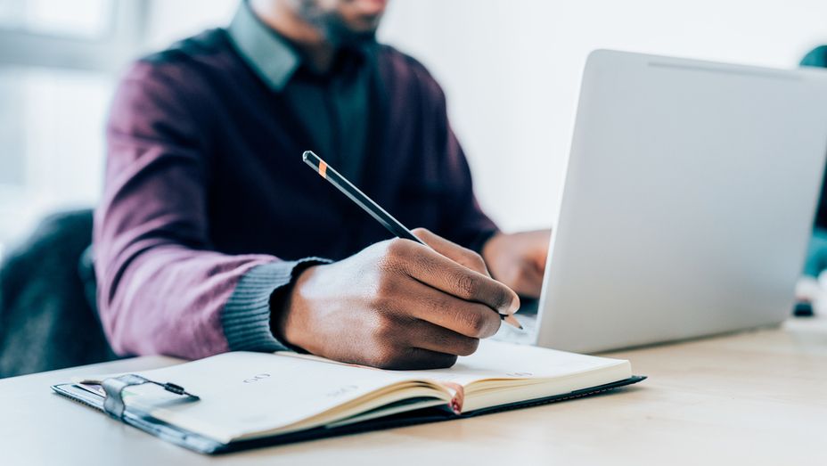 a man is writing in a notebook while using a laptop computer .