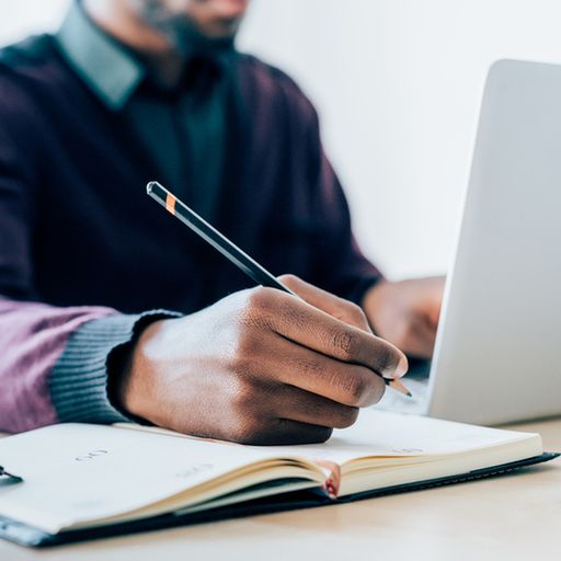 a man is writing in a notebook while using a laptop computer .