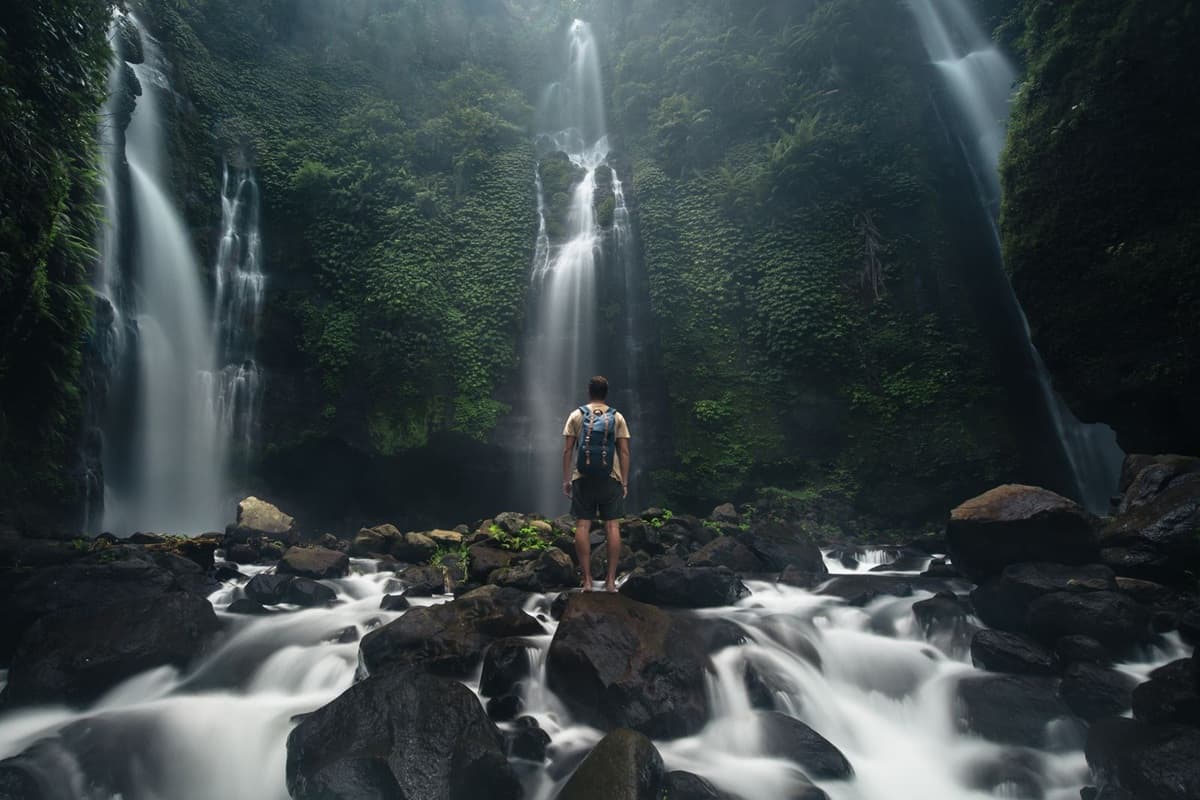 Backpacker views multiple waterfalls cascading down a lush cliff, with blurred water below.