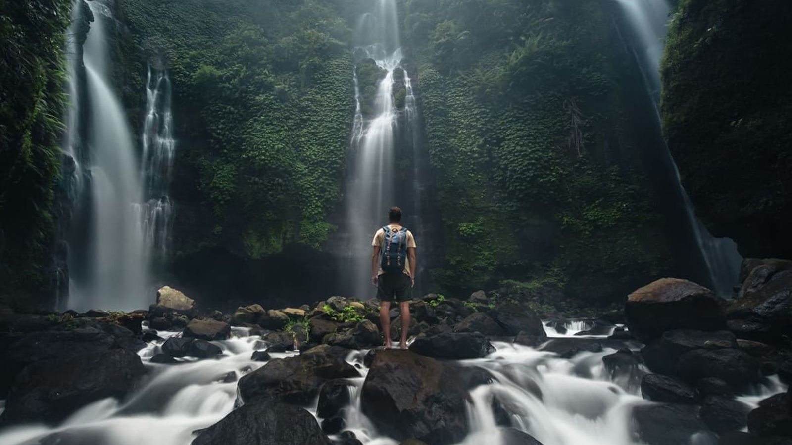 Backpacker views multiple waterfalls cascading down a lush cliff, with blurred water below.