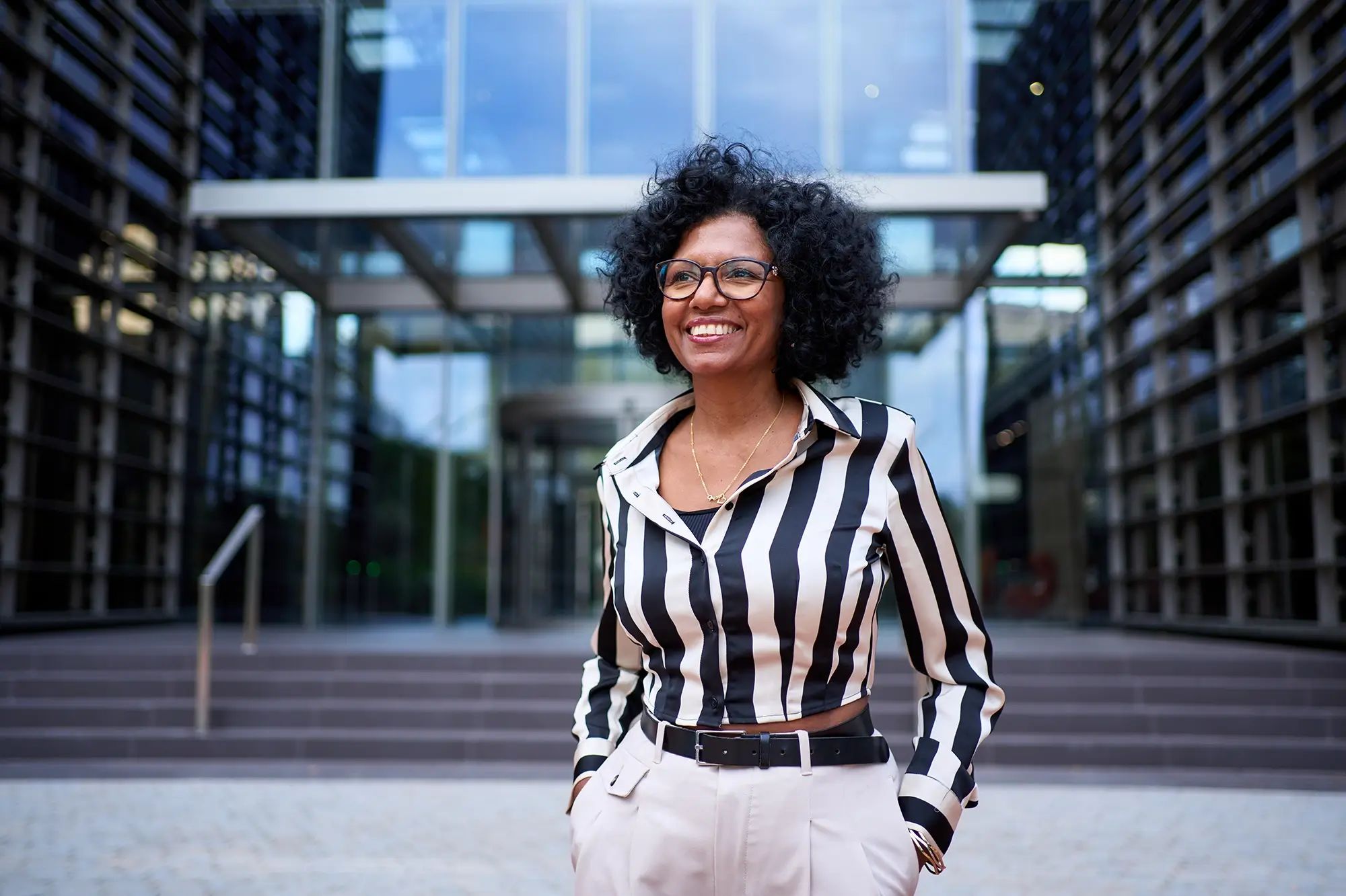 A smiling Black woman in a striped shirt and glasses stands outside a modern office building.