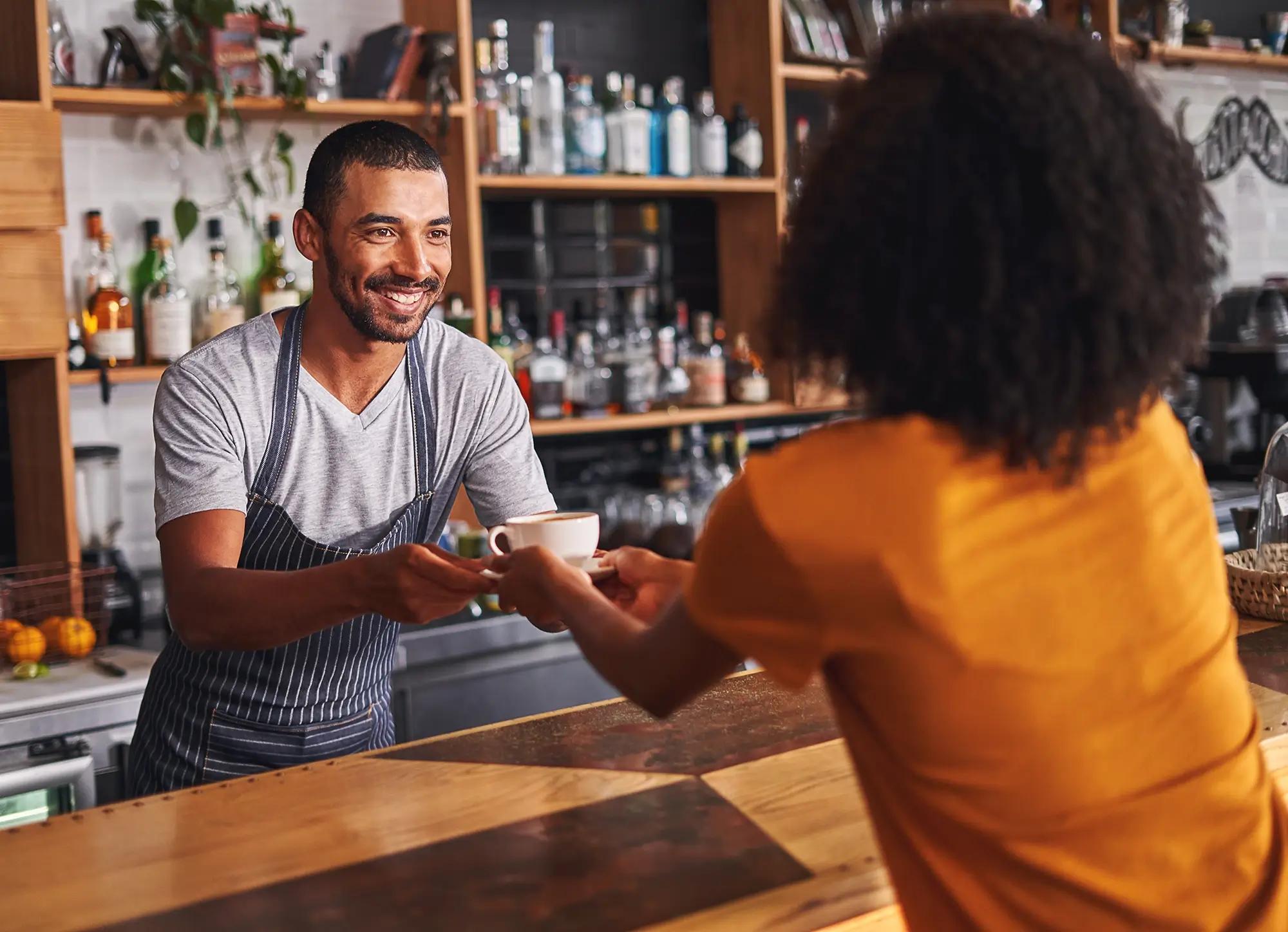 A smiling barista hands a coffee cup to a customer across a wooden counter.