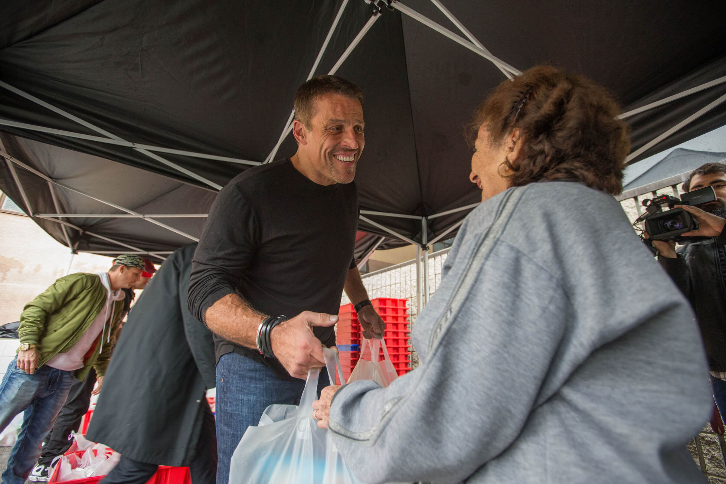Five smiling people, one man and four women, pose outdoors under blue tents; three women wear black "BEY GOOD" shirts.
