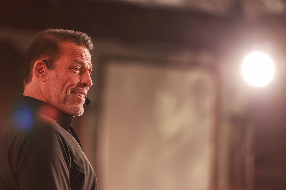 Tony Robbins smiling and wearing a headset microphone, with a bright light in the background.