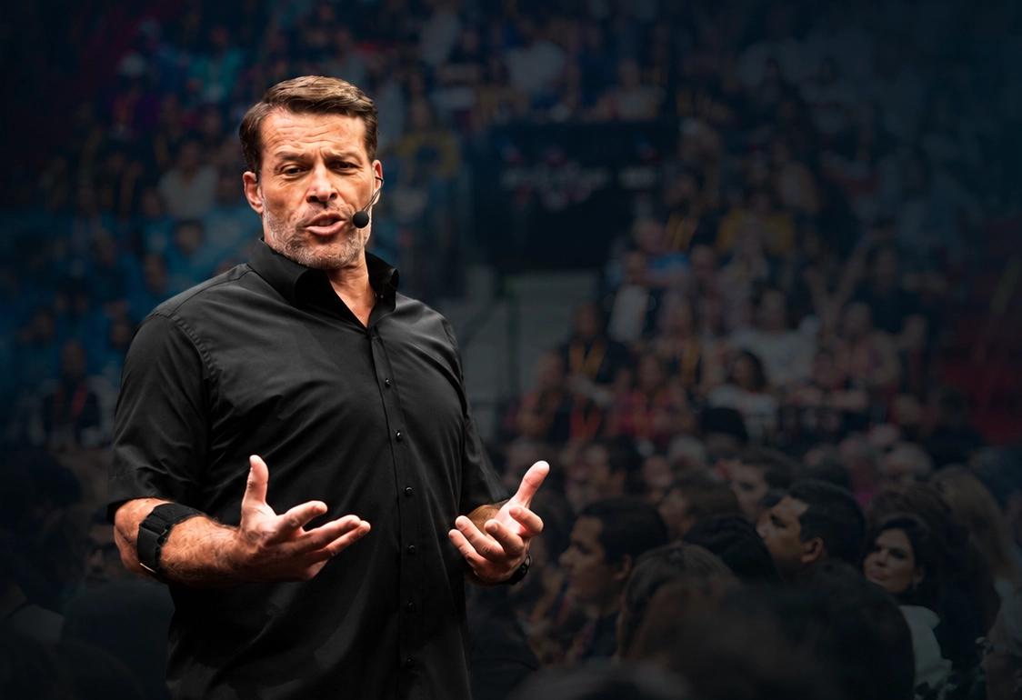 Tony Robbins speaks with a headset mic, gesturing to a blurred audience in the background.