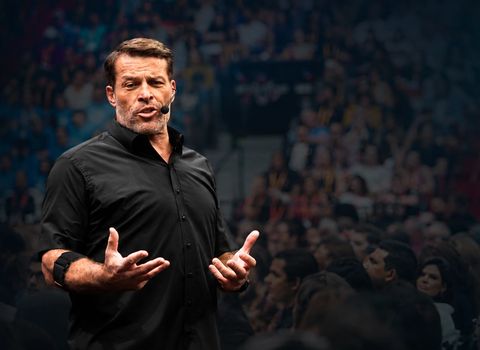 Tony Robbins speaks with a headset mic, gesturing to a blurred audience in the background.