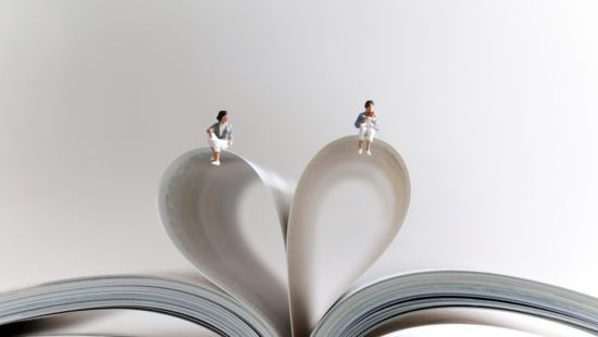 two miniature people are sitting on top of a heart shaped book .