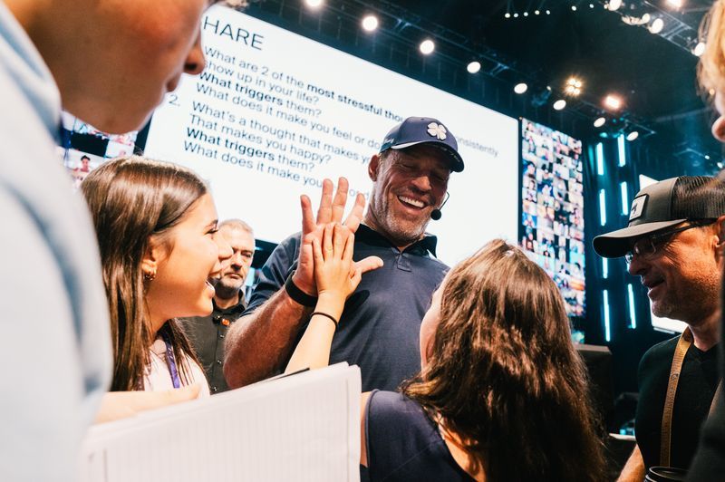 A smiling man in a baseball cap high-fives a young woman at a live event, with a large screen displaying questions in the background.