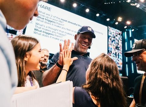 A smiling man in a baseball cap high-fives a young woman at a live event, with a large screen displaying questions in the background.