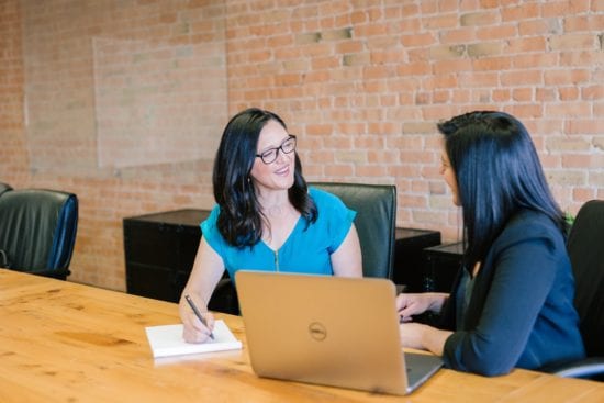 two women are sitting at a table with a laptop and talking to each other .