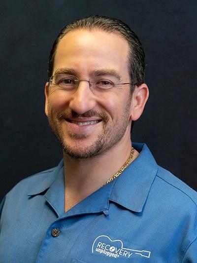 A smiling man with dark hair, glasses, and a beard wears a blue shirt with a "Recovery Unplugged" logo and a gold chain.
