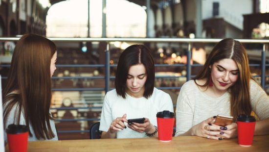 three young women are sitting at a table looking at their phones .