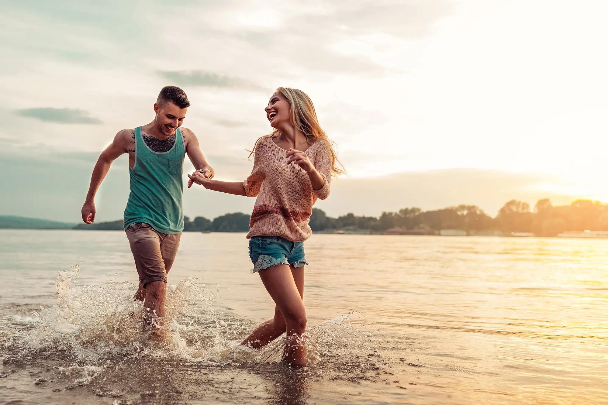 A happy couple holding hands, laughing, and running through shallow water at sunset.