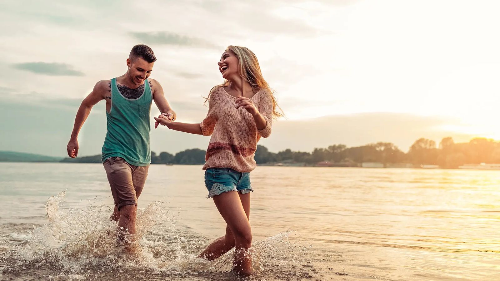 A happy couple holding hands, laughing, and running through shallow water at sunset.