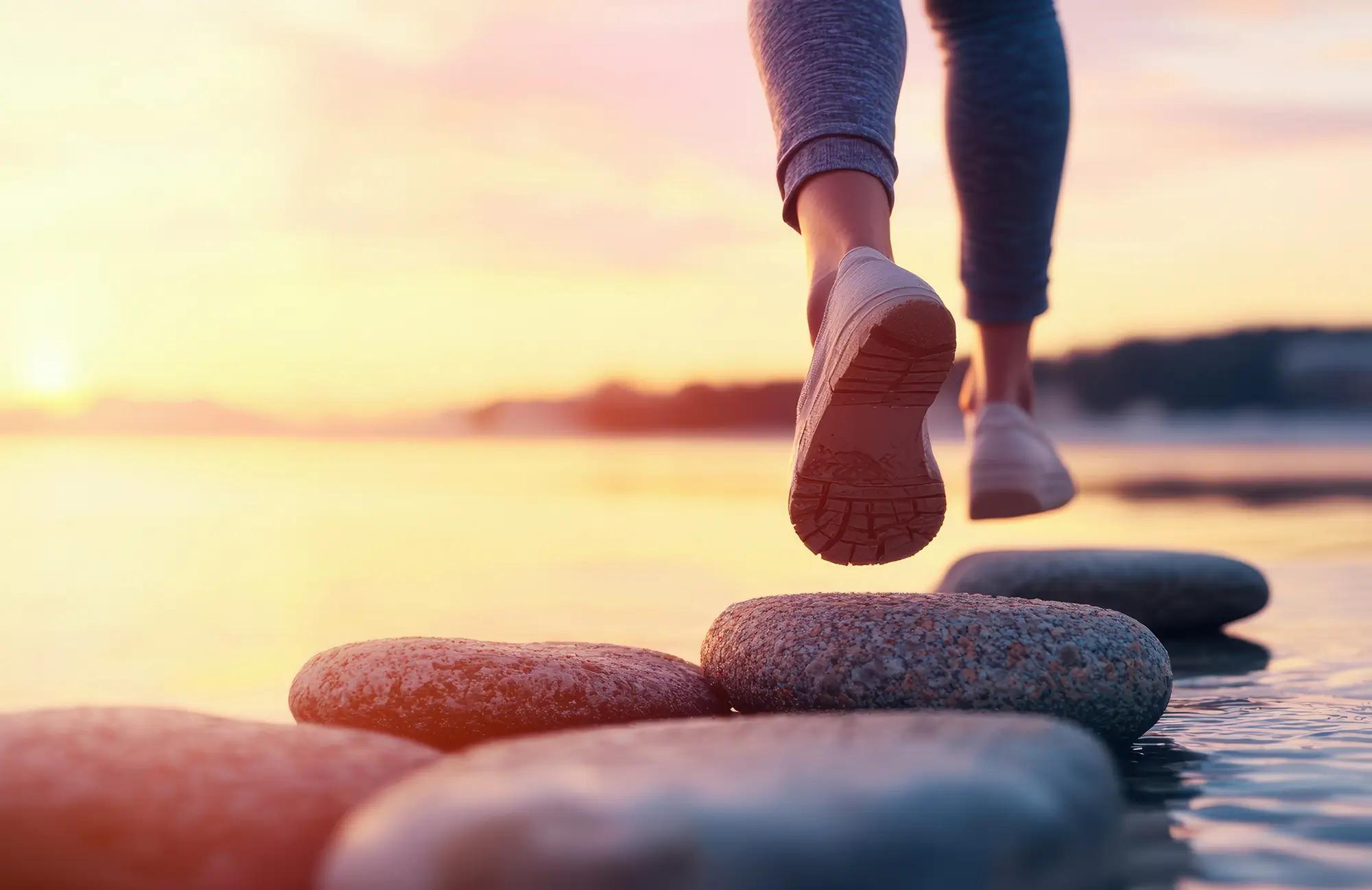 Feet in sneakers stepping on stones across water at sunset.