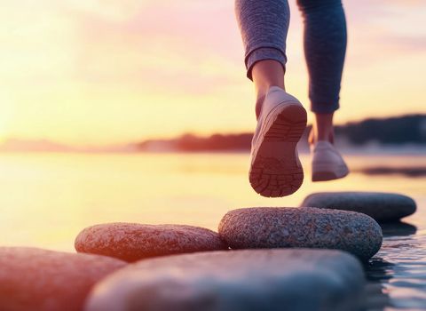 Feet in sneakers stepping on stones across water at sunset.