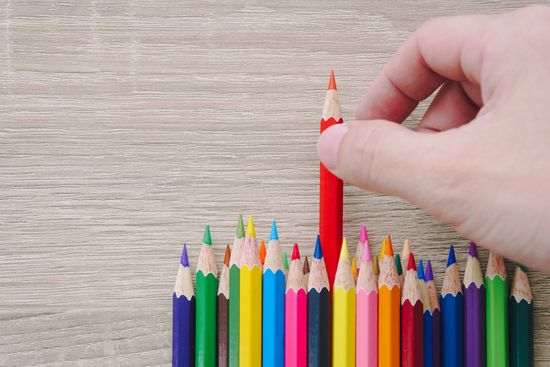 a hand is holding a red pencil in front of a row of colored pencils .