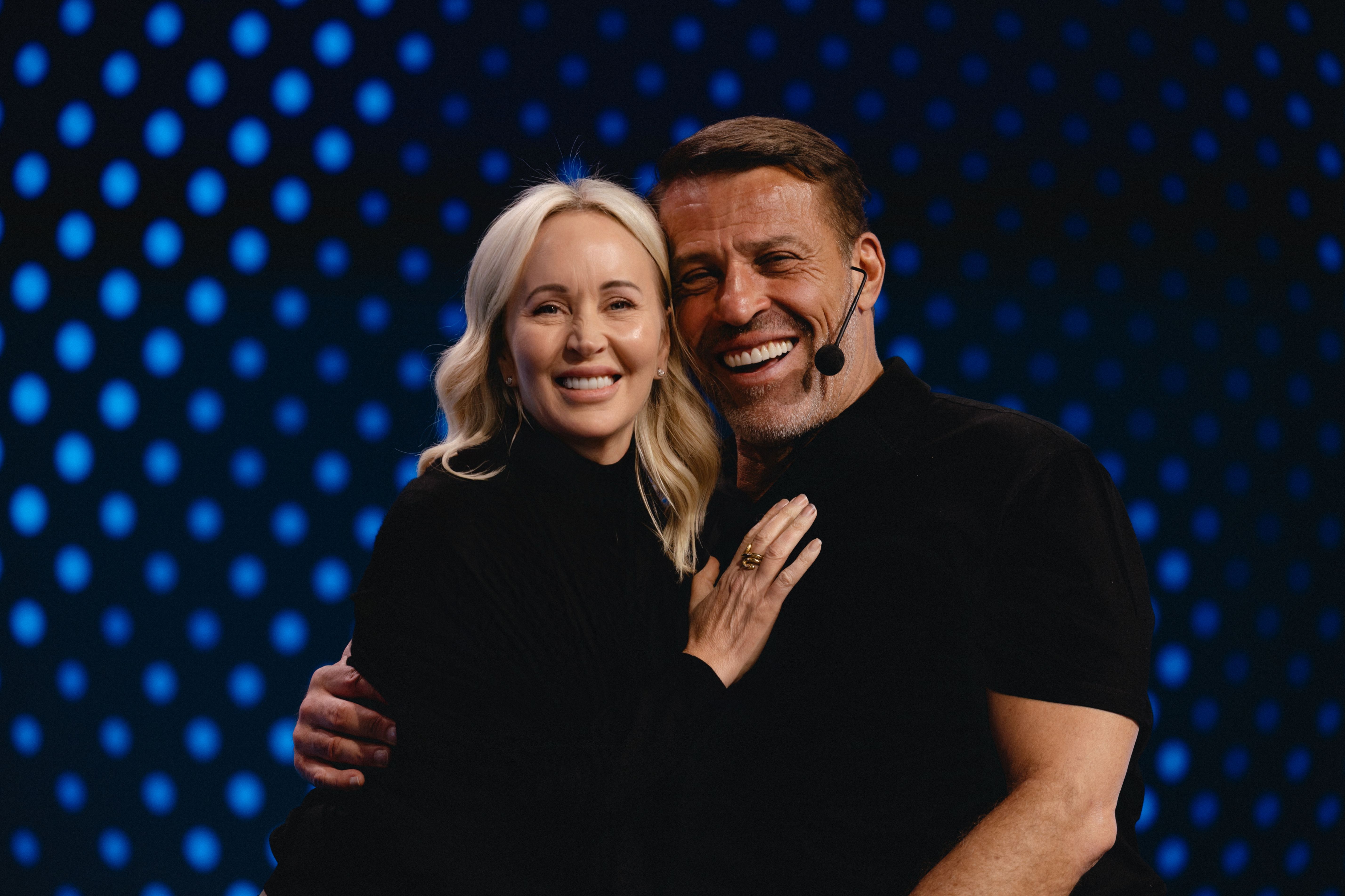Tony and Sage Robbins embracing on stage in front of a blue LED backdrop.