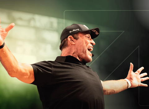 Tony Robbins energetically speaking with arms outstretched, wearing a black cap and shirt.