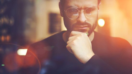 a man wearing glasses is sitting in front of a window with his hand on his chin .