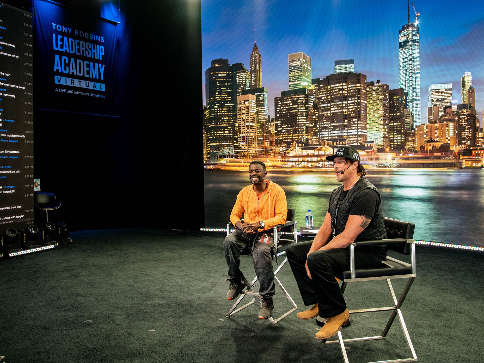 two men are sitting in director's chairs in front of a city skyline displayed on a screen